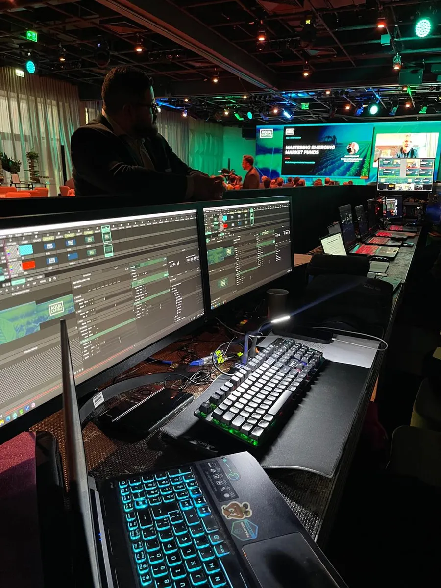 Mesa de corte com dois monitores de switcher durante a cobertura de um congresso corporativo, com o palco iluminado ao fundo